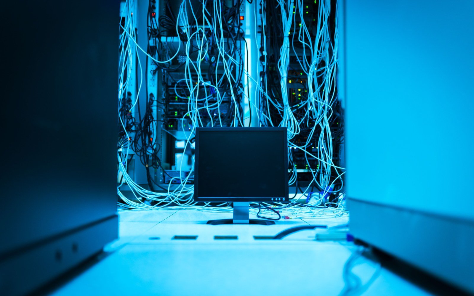 A data hall aisle seen between two cabinets, cabling hanging in loops behind a blank monitor on the raised floor