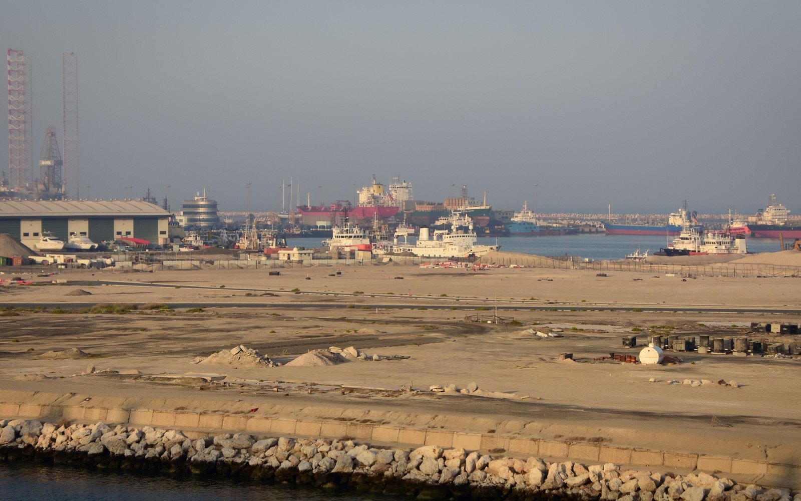 An industrial port in Dubai in flat daylight, cargo vessels and quayside sheds along the far shore behind a wide expanse of reclaimed ground and a rock breakwater