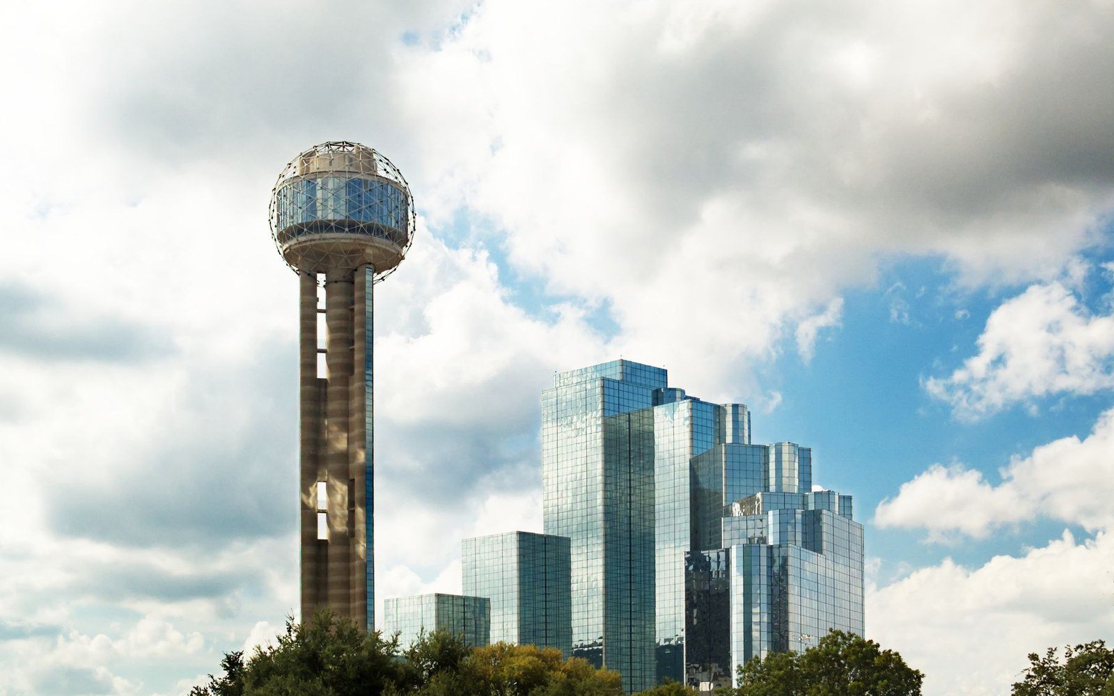 Reunion Tower and mirrored office towers rising above trees in Dallas, Texas under a cloudy sky