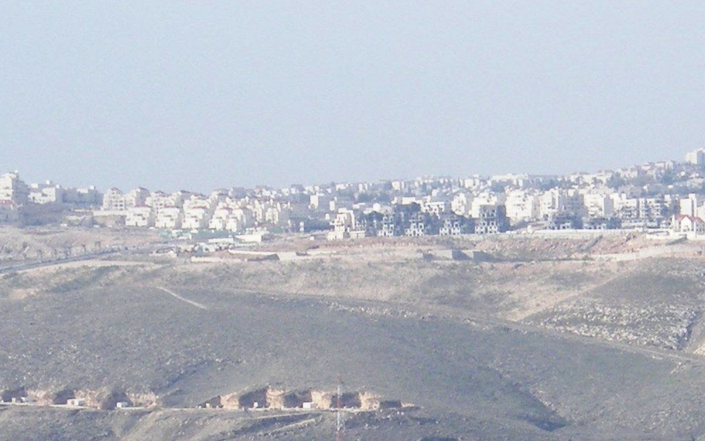 Bare stony hills in hazy light, with a band of white low-rise housing running along the ridge on the skyline and empty scrub ground falling away in front of it