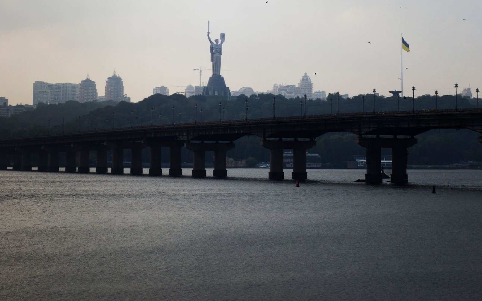 The Paton Bridge crossing the Dnipro in Kyiv under a heavy sky, with the Motherland Monument rising from the wooded bank beyond