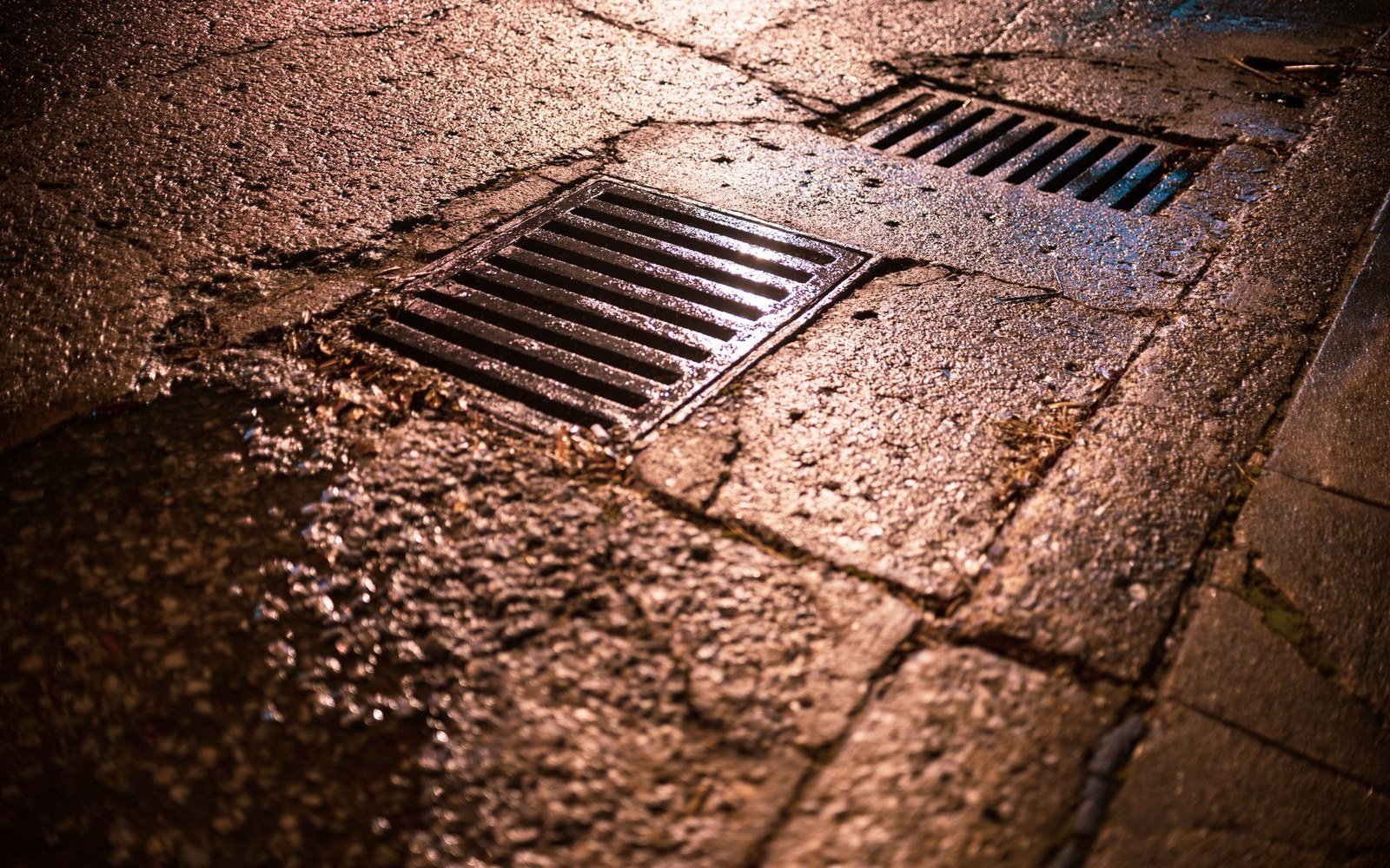 Two metal drainage grates set into a wet road surface at night, with rainwater standing on the asphalt around them