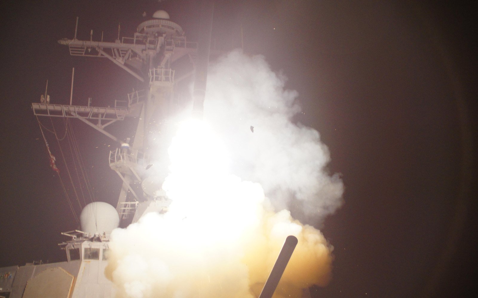 A Tomahawk missile leaving the forward launch deck of a guided missile destroyer at night, the plume lighting the mast and radar dome