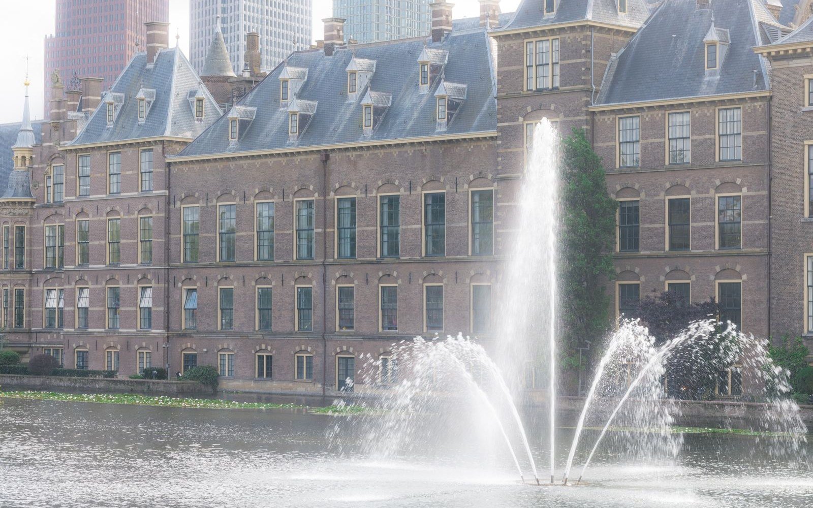 The brick and slate range of the Binnenhof in The Hague seen across its lake, with fountain jets in the foreground and modern towers rising behind the roofline