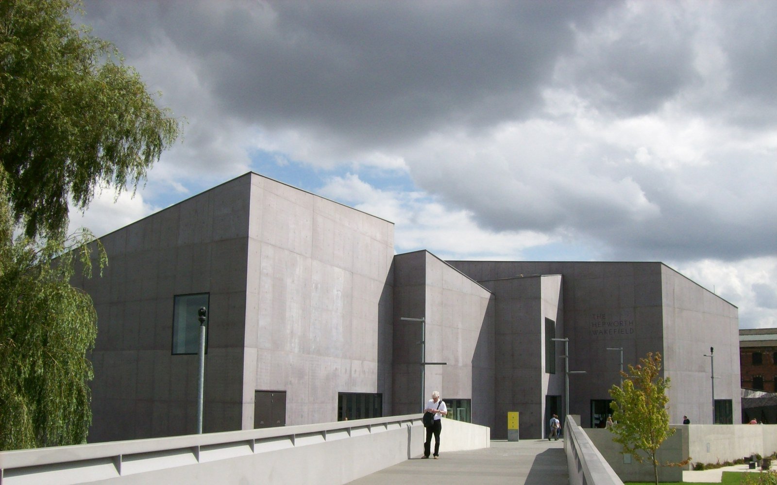 A low grey stone gallery building beside a walkway and a weeping tree under a heavy sky