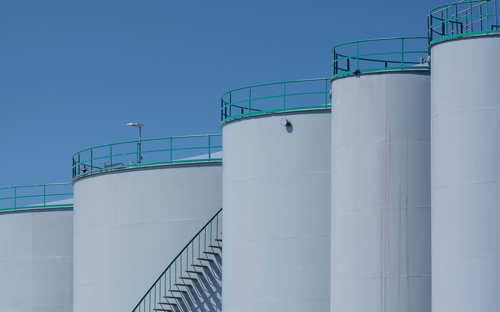 A row of white cylindrical bulk liquid storage tanks with green handrails against a clear sky
