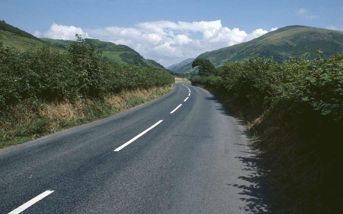 A country road curving between hedgerows below green hills in the Welsh countryside