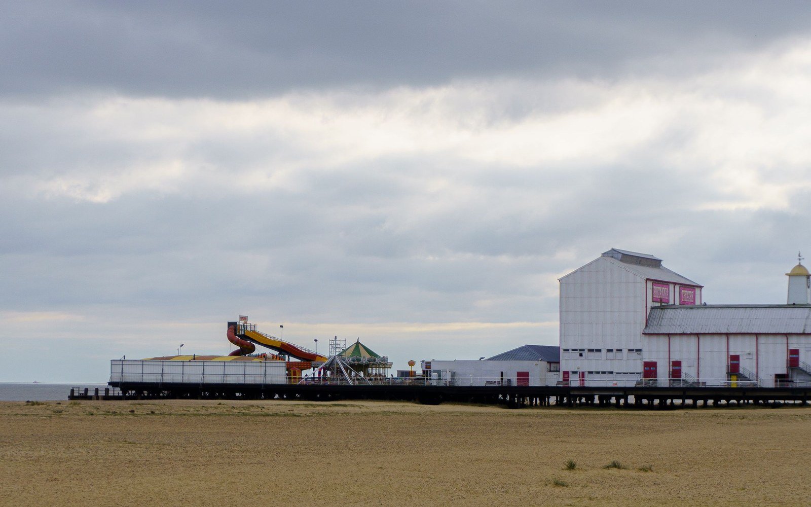 A seaside pleasure pier carrying rides and pavilions seen from a wide empty beach under a grey sky
