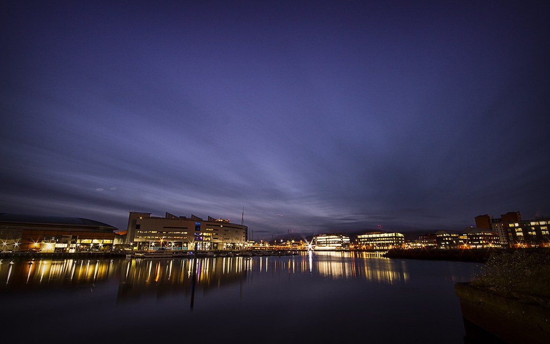 The waterfront at Titanic Quarter in Belfast at dusk, with lit buildings reflected in still water