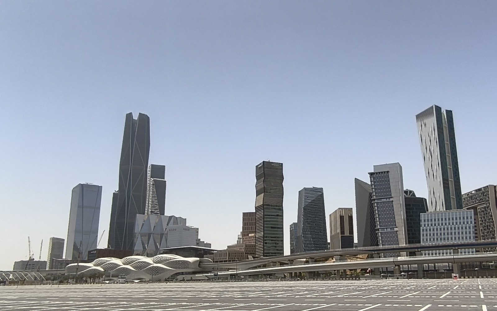 Towers of the King Abdullah Financial District in Riyadh stand behind an elevated railway and a domed station canopy across an empty surface car park in daylight