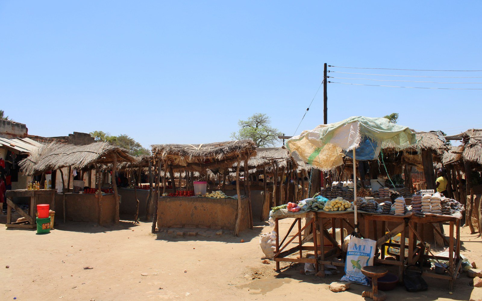 A rural market in Zambia, a line of thatched stalls on bare ground with produce and bottled oil set out on timber tables