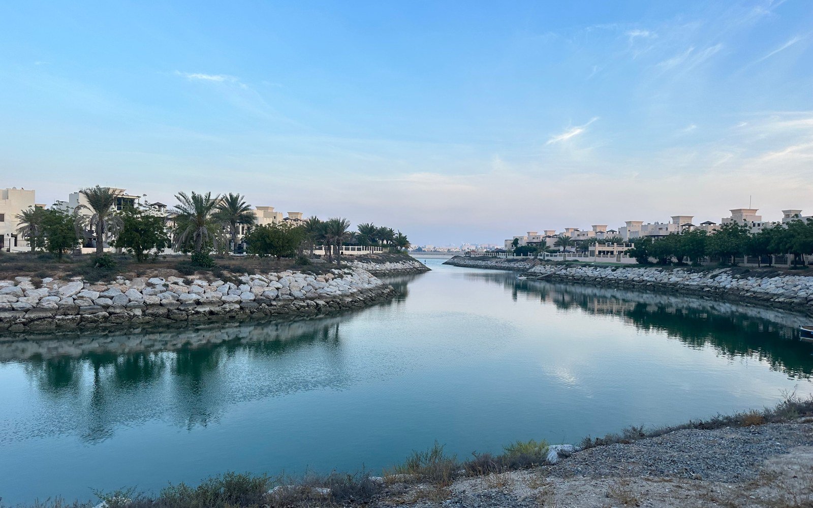 A still water channel between two rock armoured banks in a residential district of Ras Al Khaimah, low white villas and palm trees on either side, the open sea at the far end under a pale evening sky