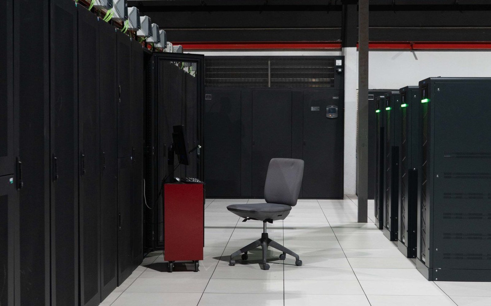 An aisle between rows of black server cabinets in a data hall, indicator lights along the racks and a pale tiled floor, no people in shot