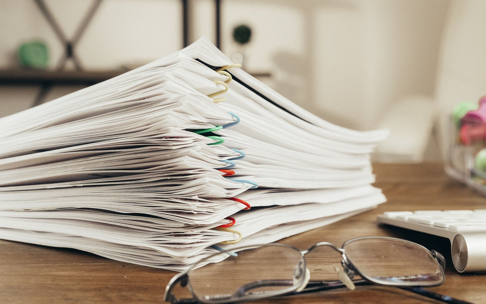 A thick stack of paper clipped in coloured bundles on a wooden desk, a pair of reading glasses in front of it and the edge of a keyboard behind