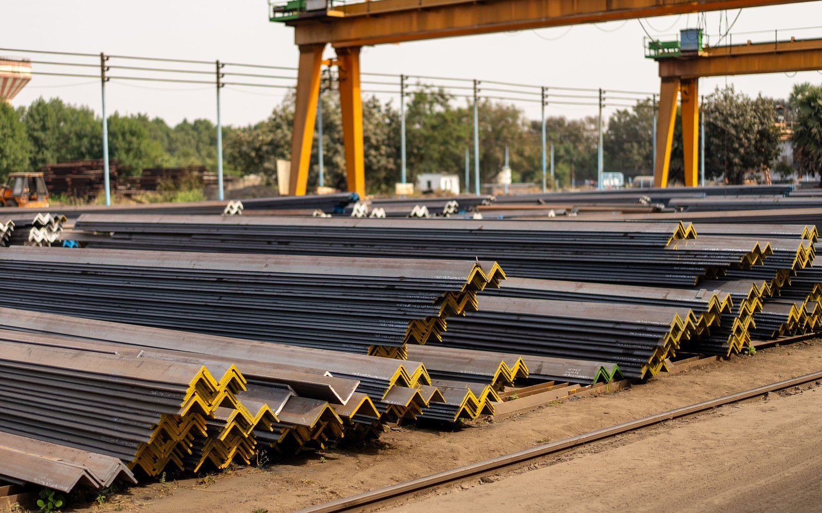 Long steel angle sections stacked in tiers in an open yard, supported on timber bearers under a yellow gantry crane, with trees and overhead lines beyond