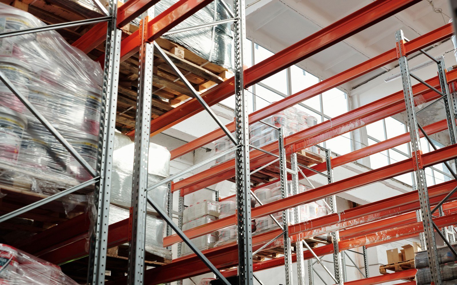 Pallet racking inside a warehouse, with galvanised uprights and orange beams carrying timber pallets and shrink wrapped loads on several levels, seen from below against a lit roof