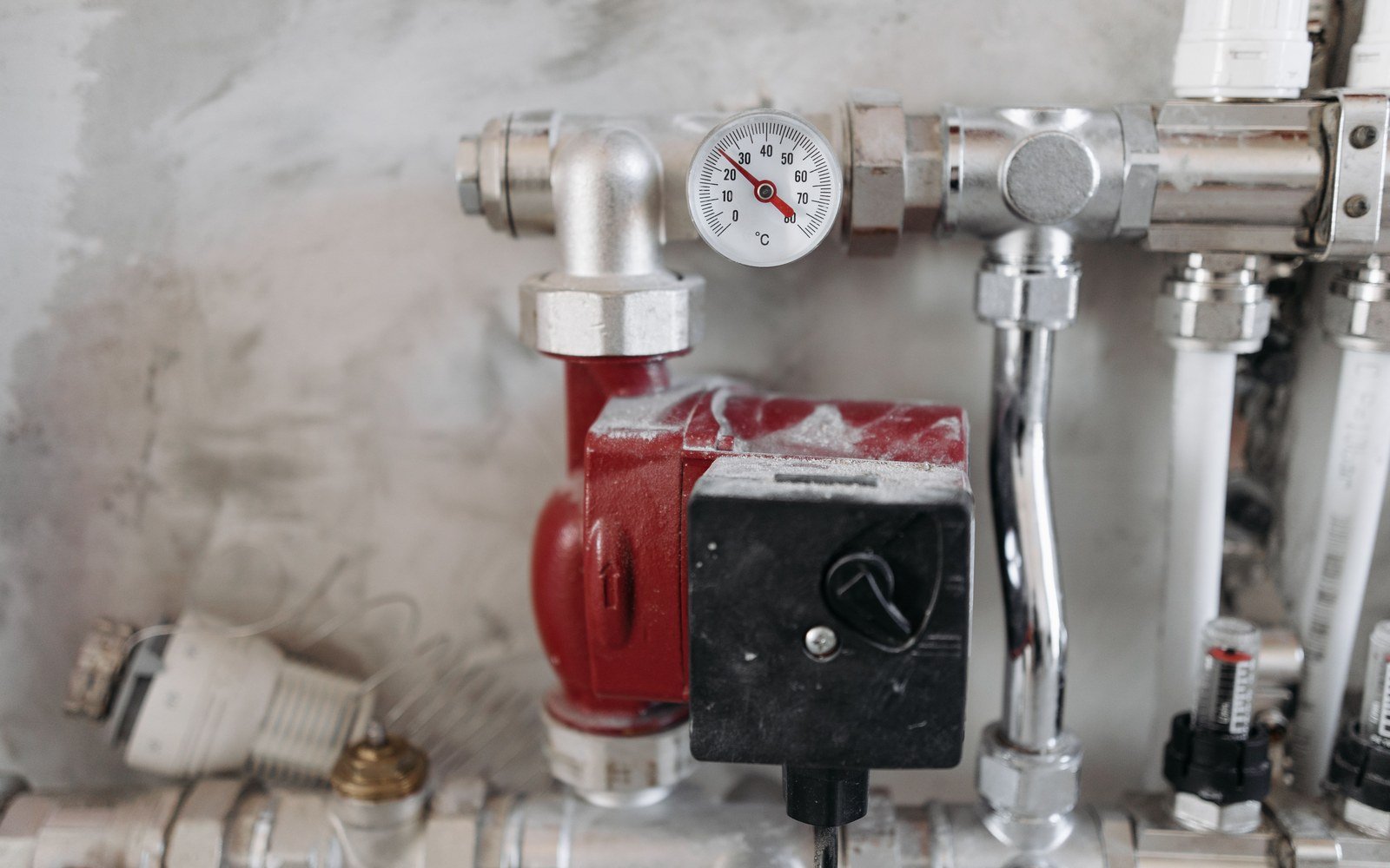 Plant room pipework in close view, a red circulating pump and a round temperature gauge mounted among chrome and white pipes against a grey wall