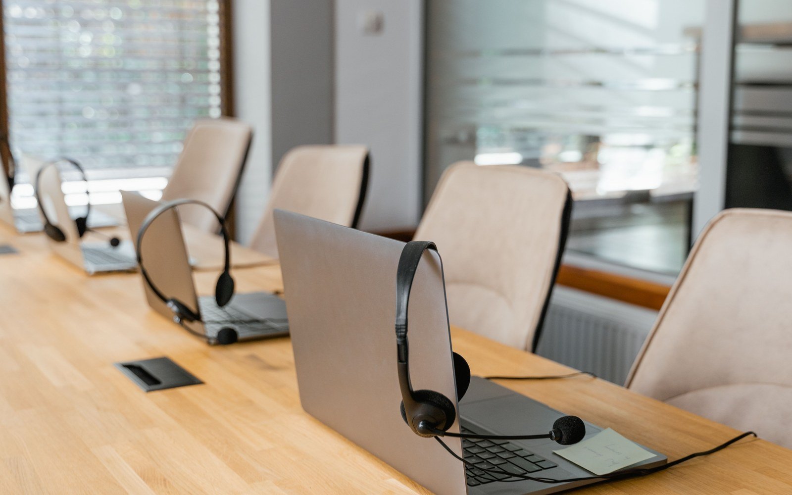 A row of open laptops with headsets resting on the keyboards along a light wooden table in an empty office