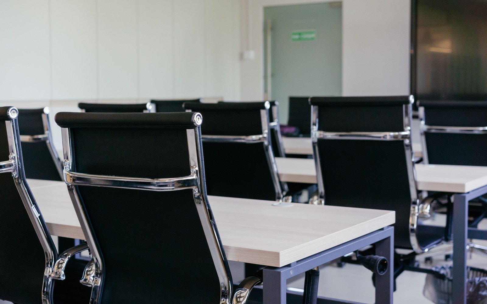 Rows of black upholstered chairs pulled up to pale conference tables in an empty meeting room, a blank display panel on the far wall