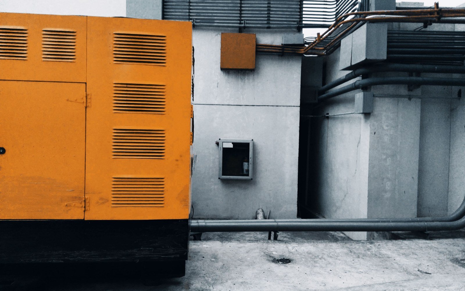 An orange enclosed generator set standing against a bare concrete wall in a service yard, louvred vents along its side panels and hinged access doors, exposed pipework running along the wall behind it