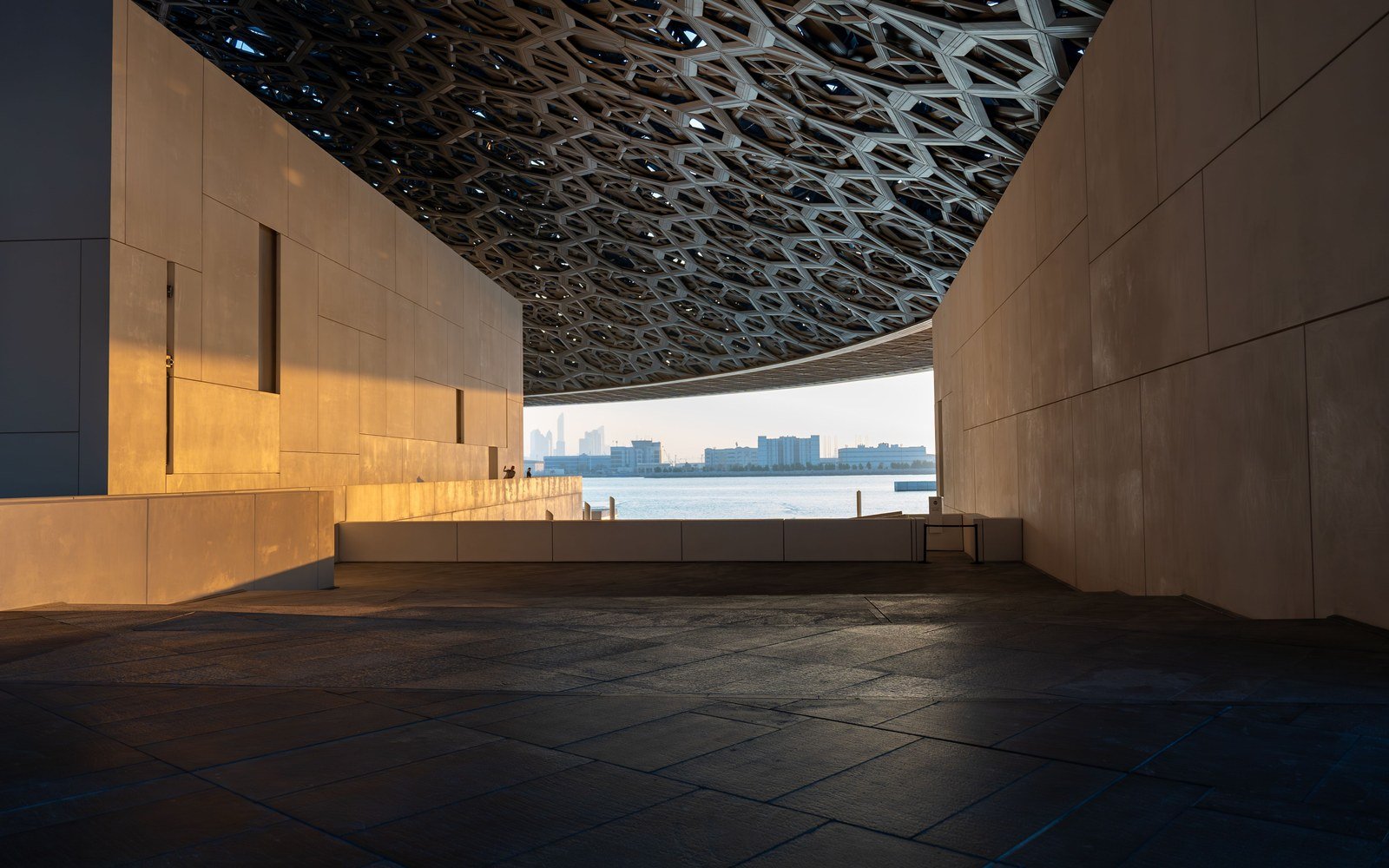 The perforated steel canopy of the Louvre Abu Dhabi seen from beneath at golden hour, pale stone walls to either side, the shadowed plaza in the foreground and a strip of sea and the city skyline framed at the far end