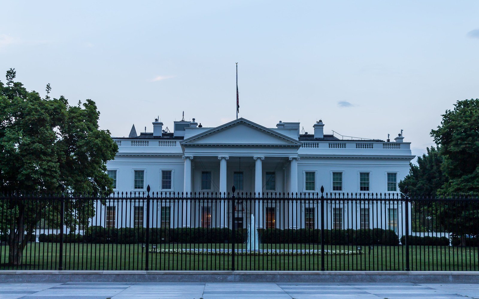 The north front of the White House in Washington behind its black iron railings, a fountain playing on the lawn