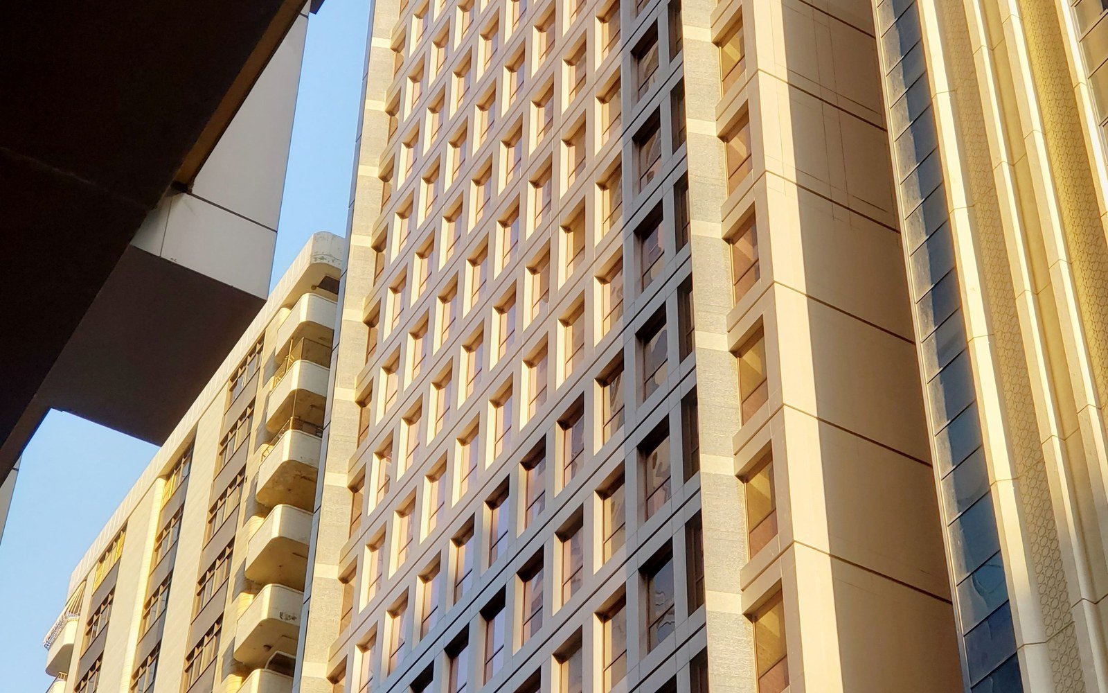 Modern residential and office towers in Abu Dhabi seen from street level in late afternoon light, a run of regular window bays on the nearest tower and balconies on the block behind, no people in the frame