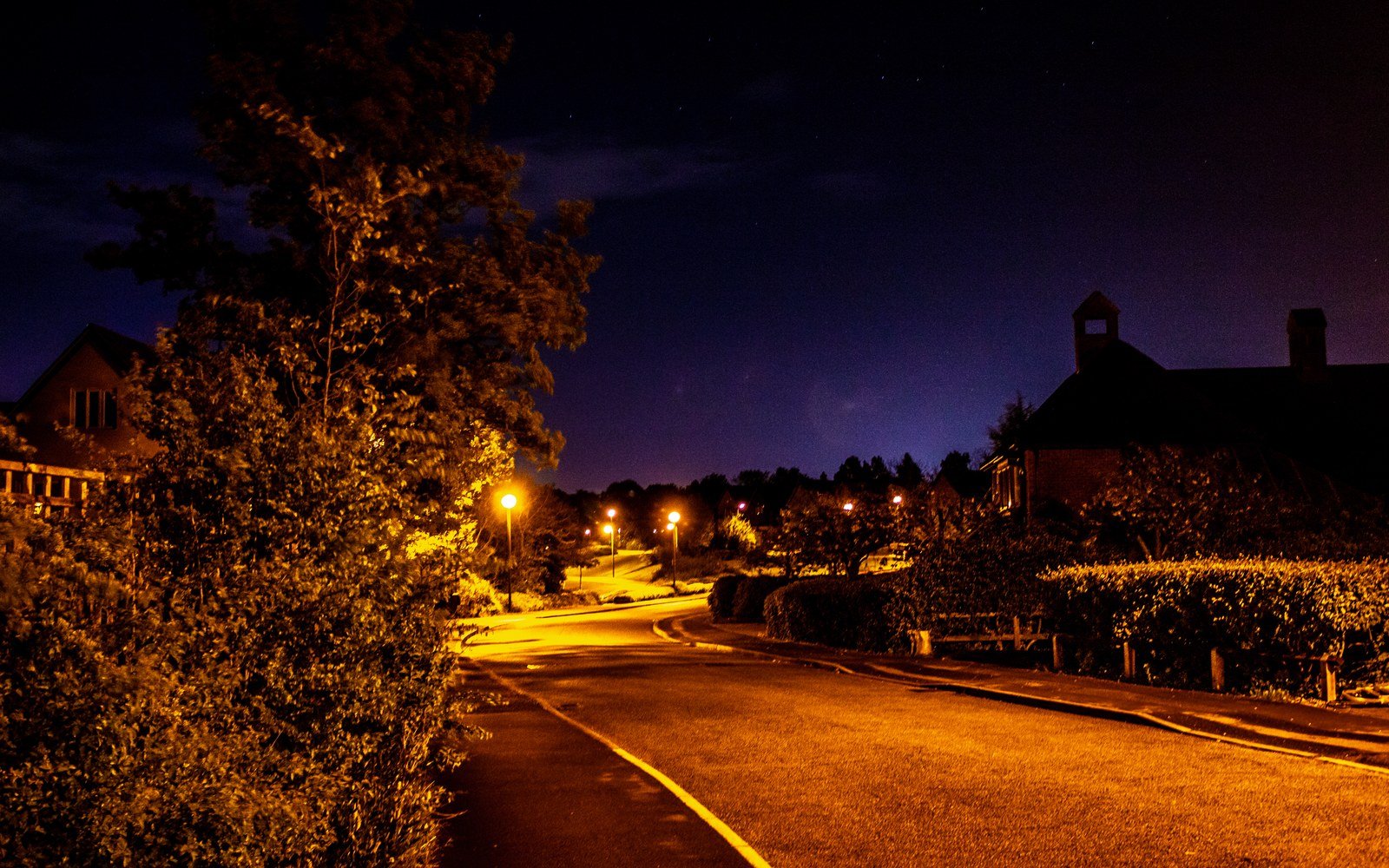 A quiet residential road at night under orange street lighting, with hedges and house roofs against a deep blue sky