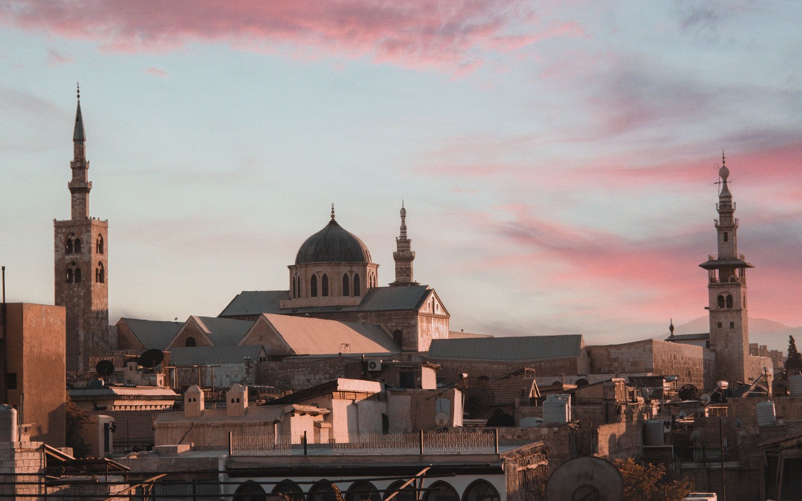 The skyline of an old city at dusk under a pink and grey sky, with a stone dome, three minarets and pitched metal roofs rising above flat rooftops