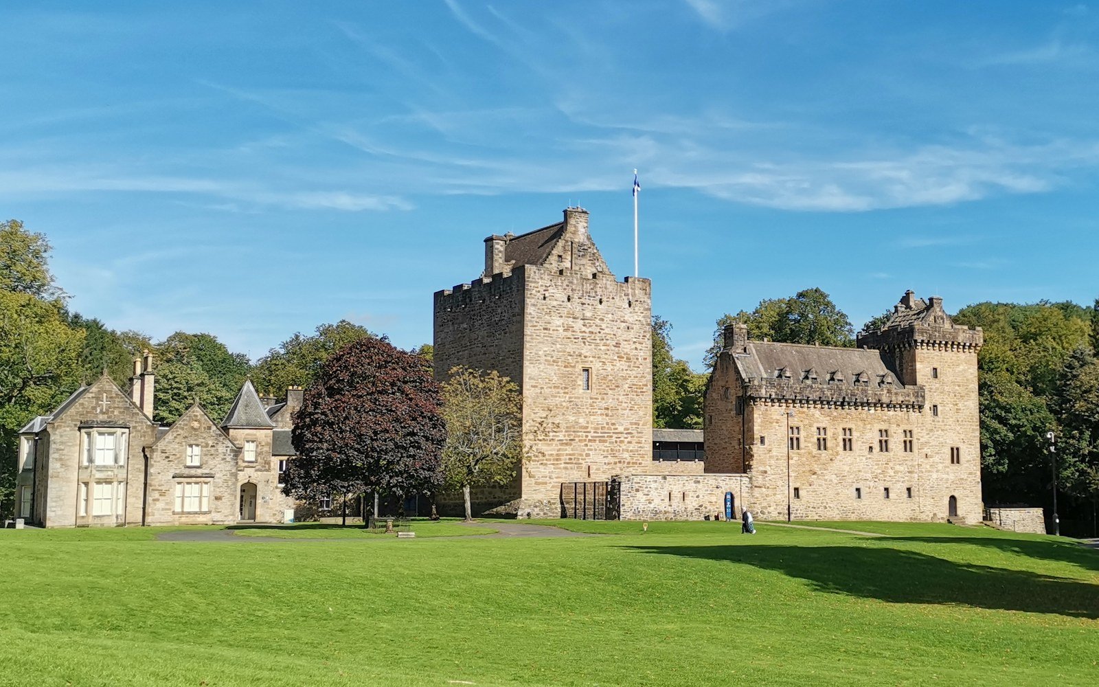 A restored stone castle with a battlemented keep and a lower range, standing on mown parkland under a clear sky