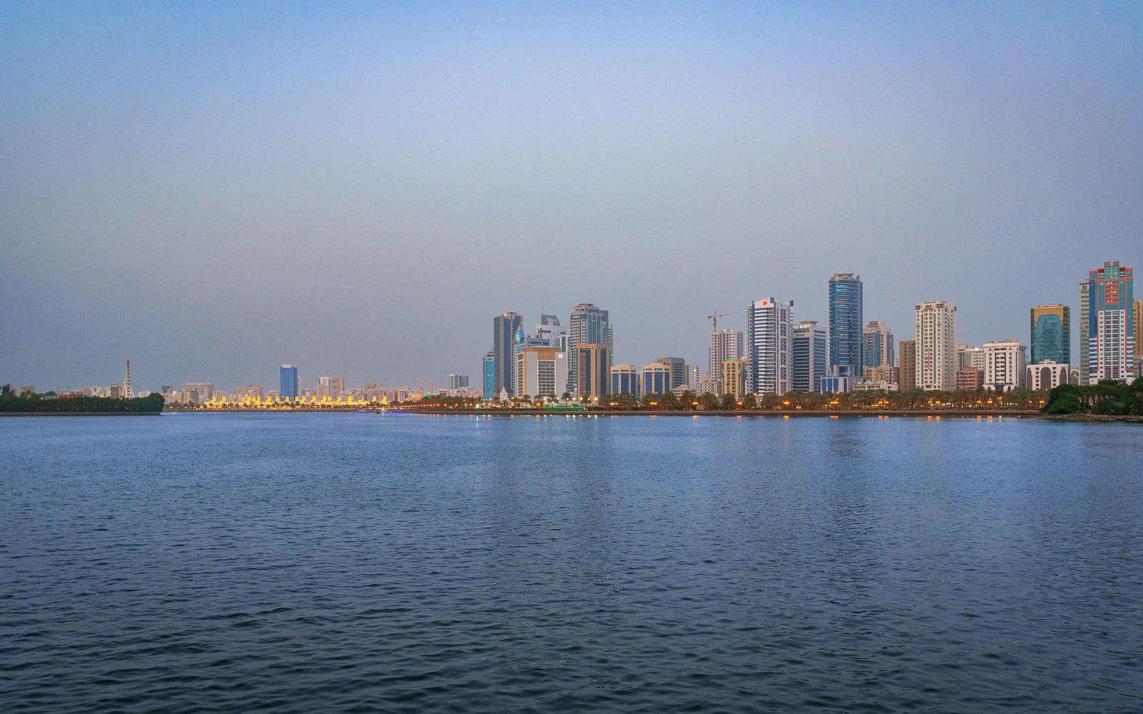 The Sharjah skyline of high-rise towers seen across calm water at dusk, the buildings reflected in the surface under a pale sky