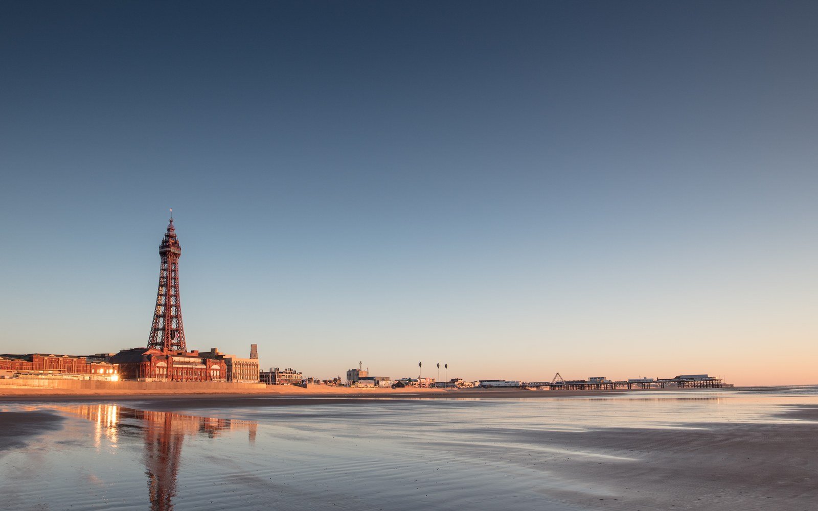 The Blackpool seafront at sunset, with the tower and the piers seen along the shoreline across wet sand