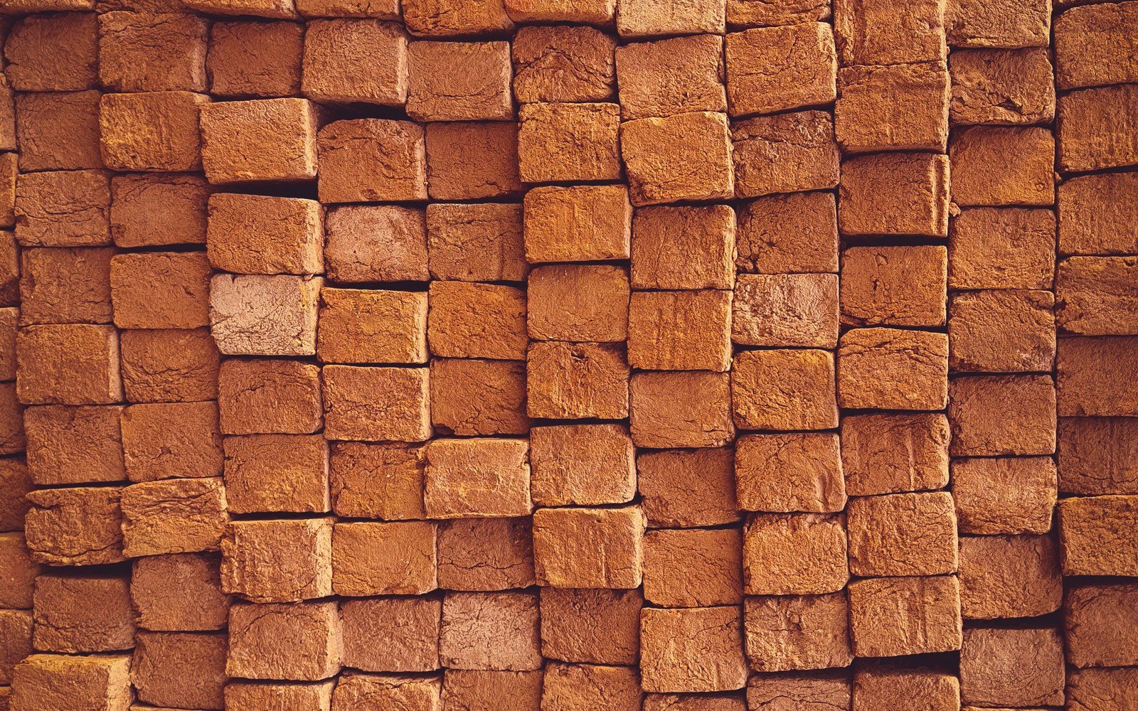 Rows of clay bricks stacked face to face in a stepped pile, filling the frame, their faces showing a rough sanded texture in warm orange and brown