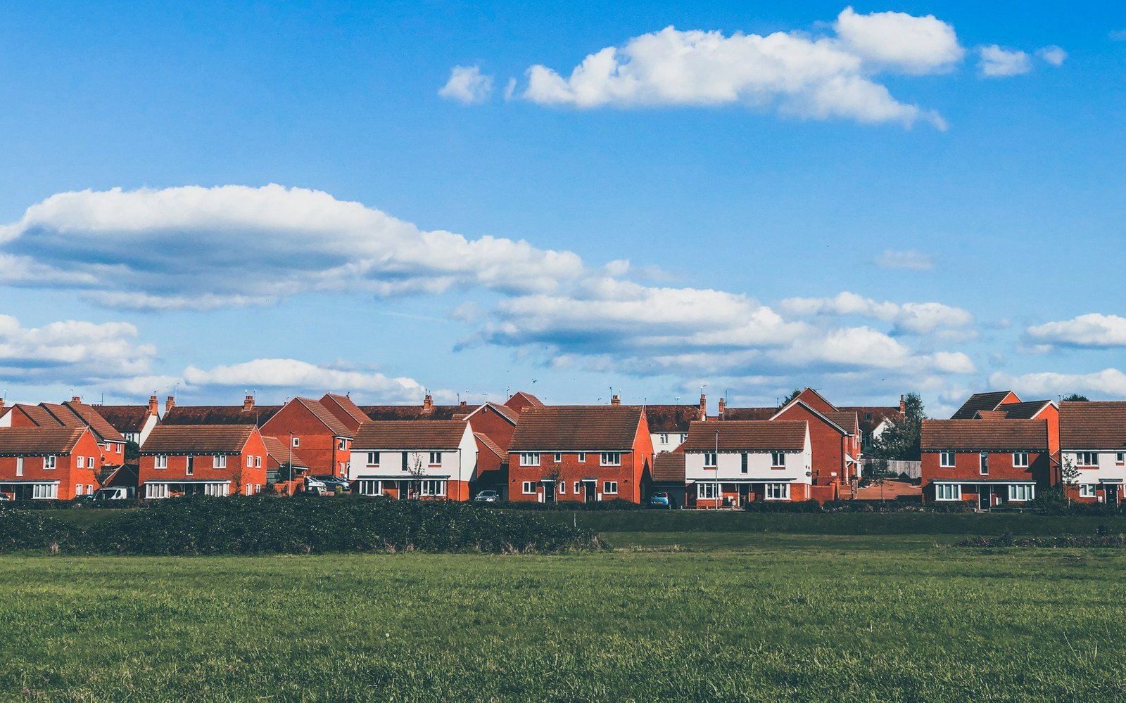 A row of modern brick and rendered houses with pitched tiled roofs standing behind a clipped hedge, seen across an open field of mown grass under a bright sky