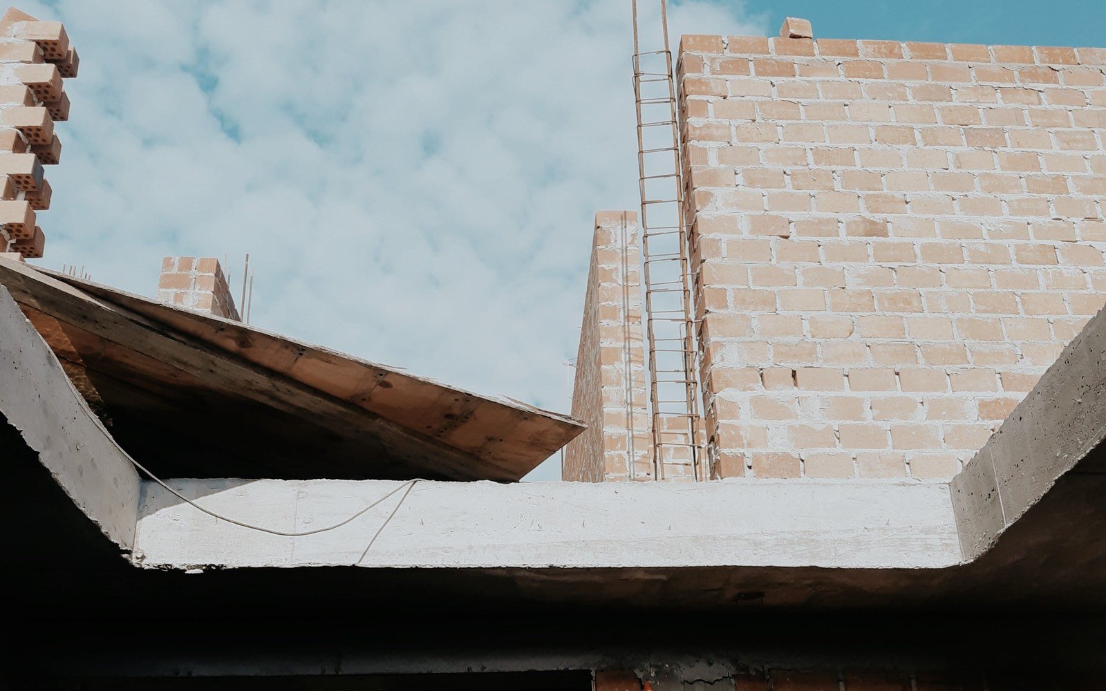 The unfinished shell of a house seen from below, brick walls open to the sky, a concrete lintel across the front and stacked bricks on the wall head