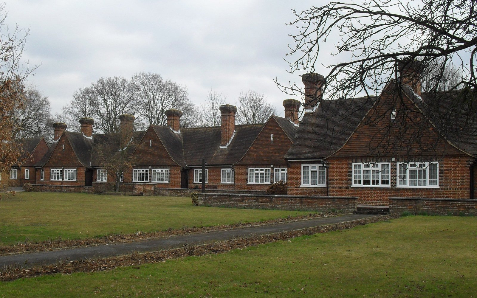 A long terrace of red brick almshouses with tall chimneys and dormer gables, set behind a lawn and a paved path