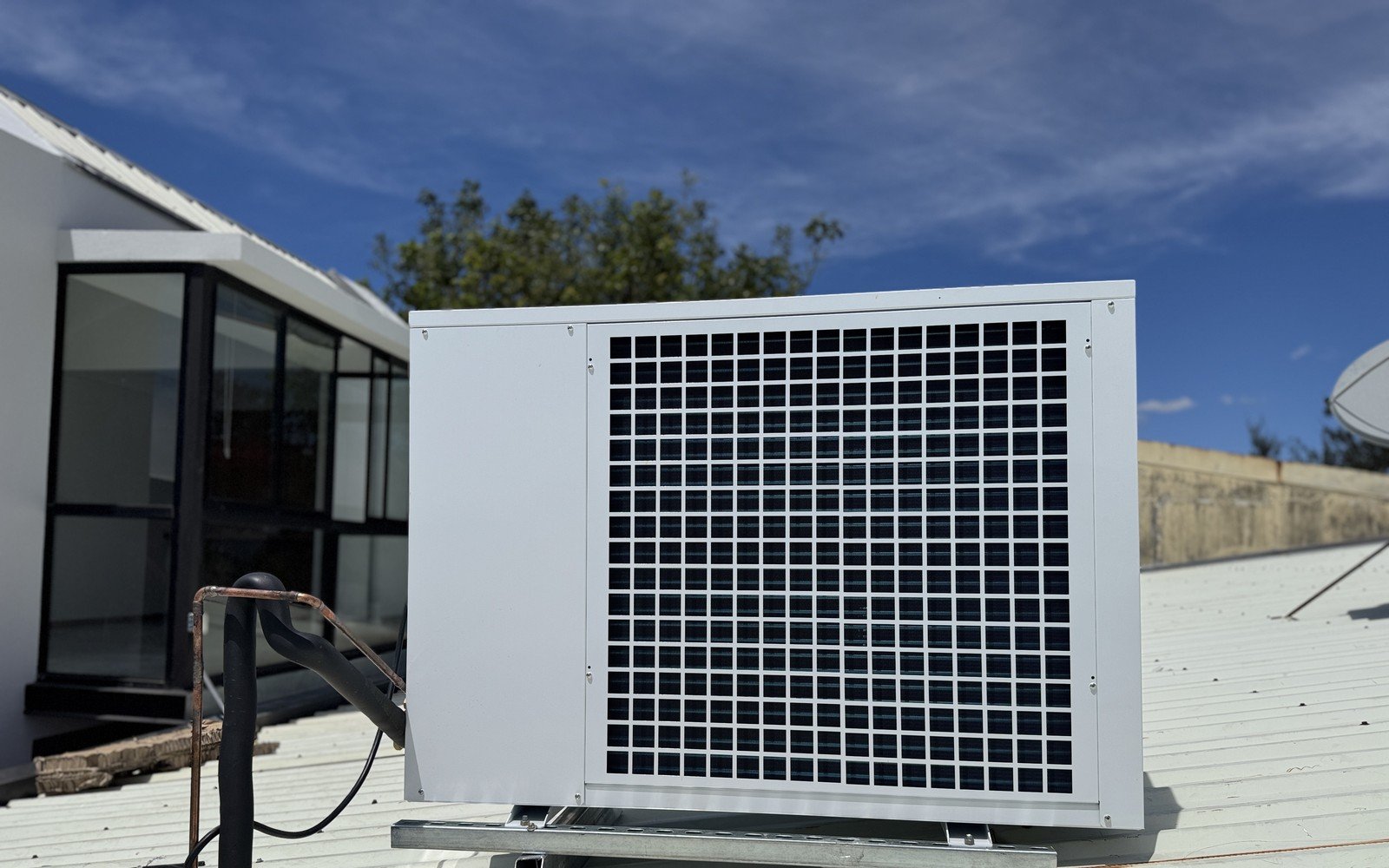 A large white air conditioning condenser standing on a galvanised frame on a corrugated metal roof, with insulated pipework running to it and a glazed single storey building behind