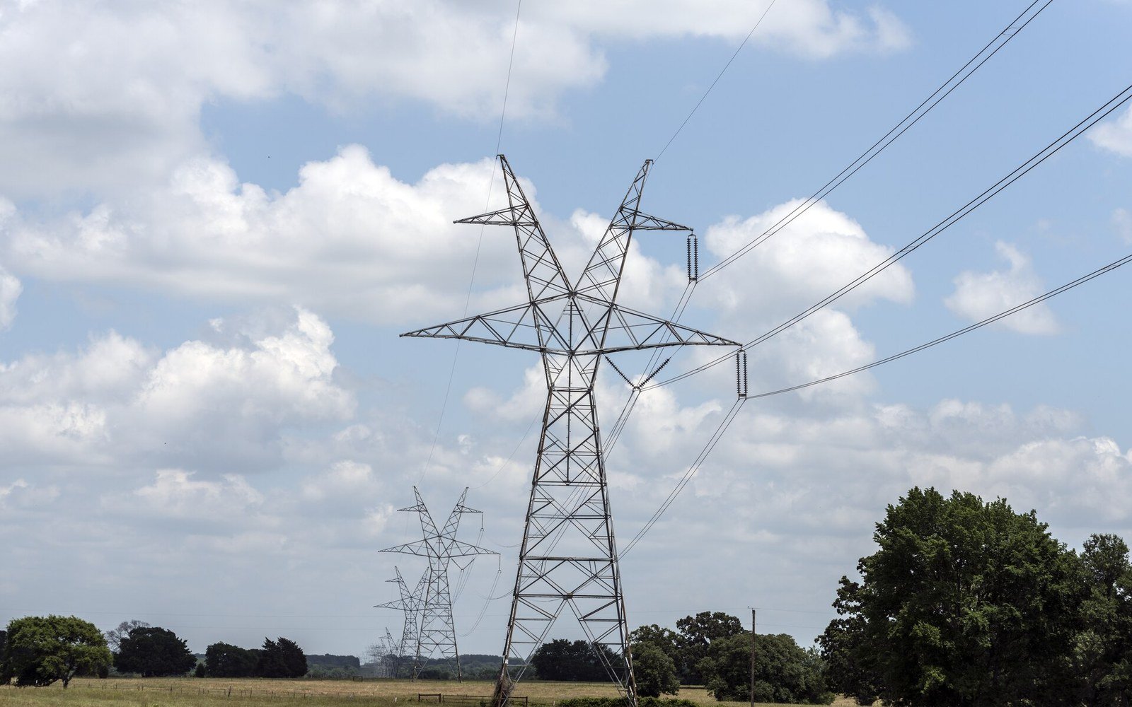 A line of steel electricity transmission towers carrying conductors across open grazing land in northeast Texas, with further towers receding towards the horizon