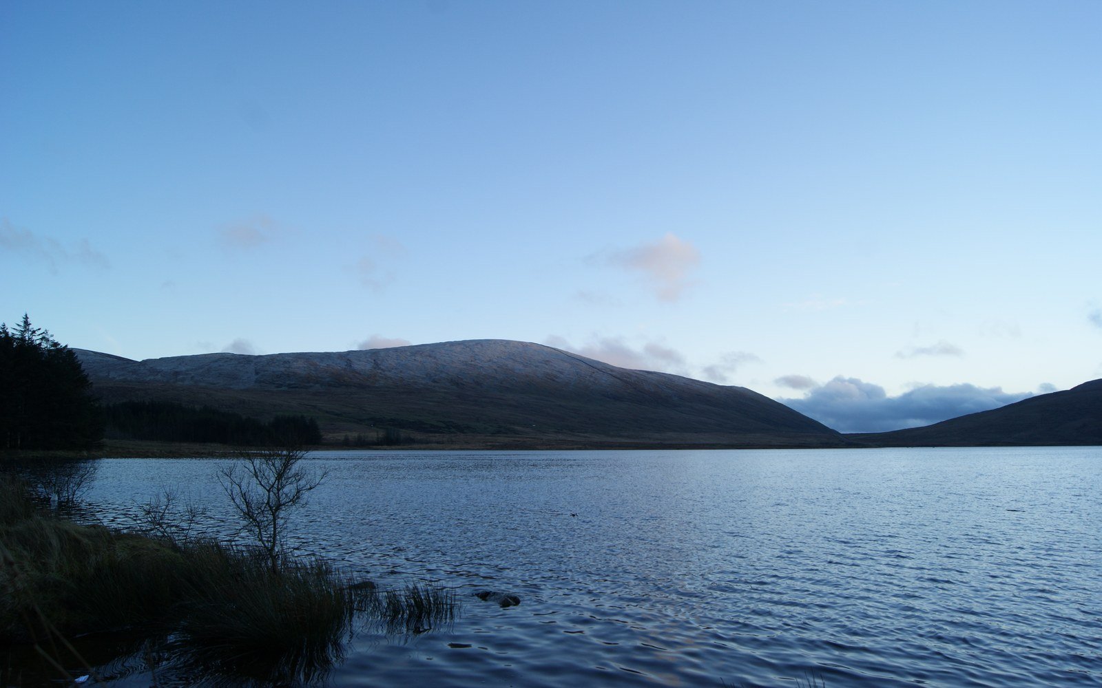 A wide upland reservoir at dusk with reeds along the near shore and a bare rounded mountain ridge closing the far end