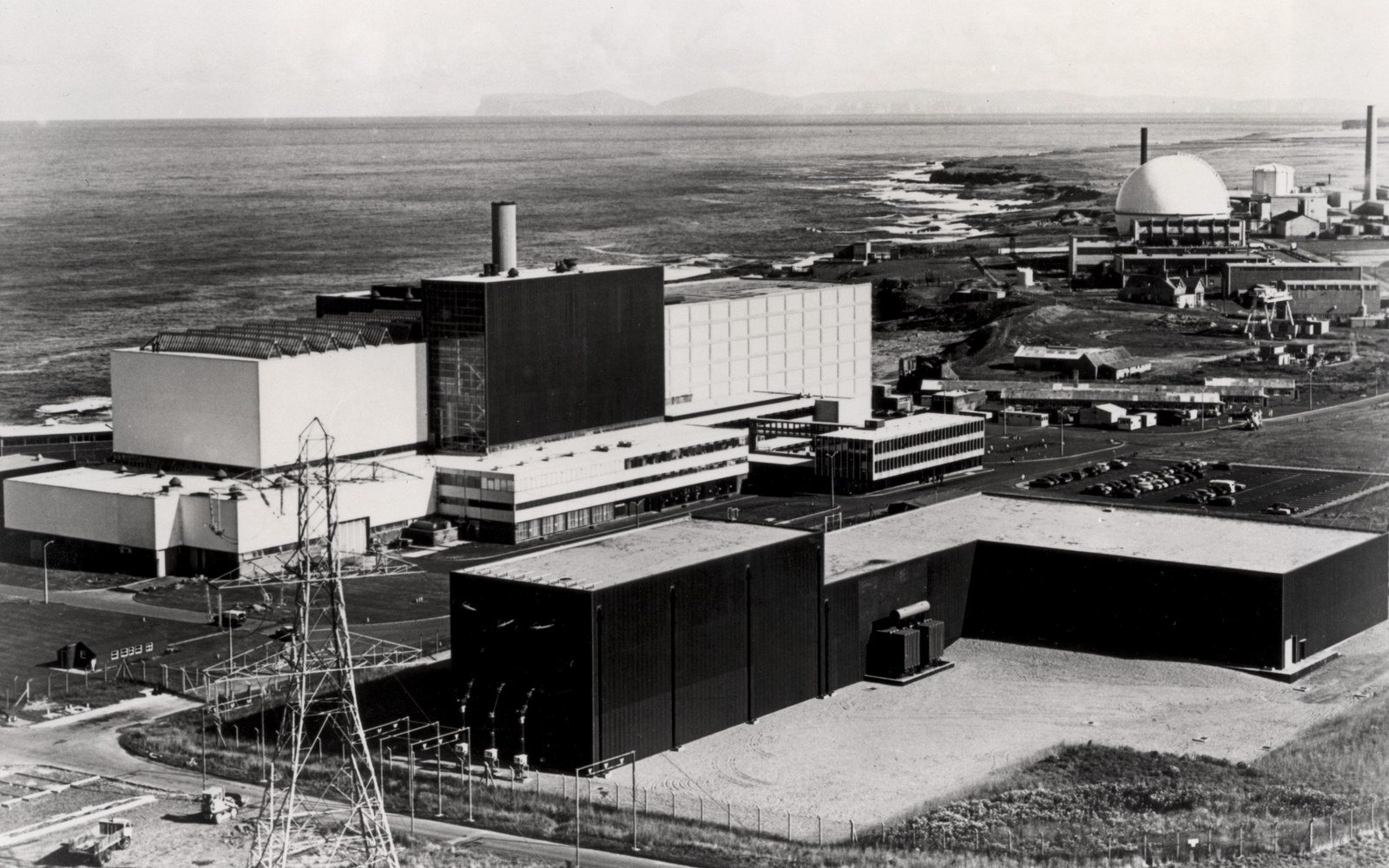 A black and white aerial photograph of a nuclear reactor complex on a flat coastal site, with a domed sphere beyond and the sea behind