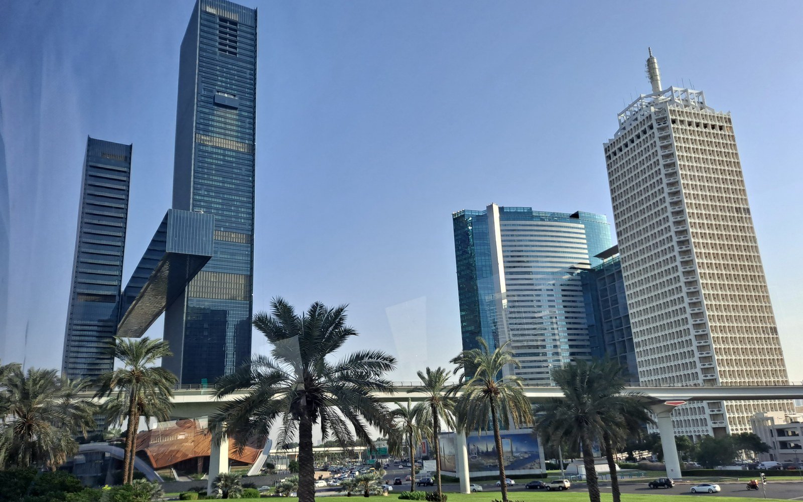 Glass and concrete towers along Sheikh Zayed Road in Dubai, with the raised metro viaduct and a row of date palms in front of them under a clear sky