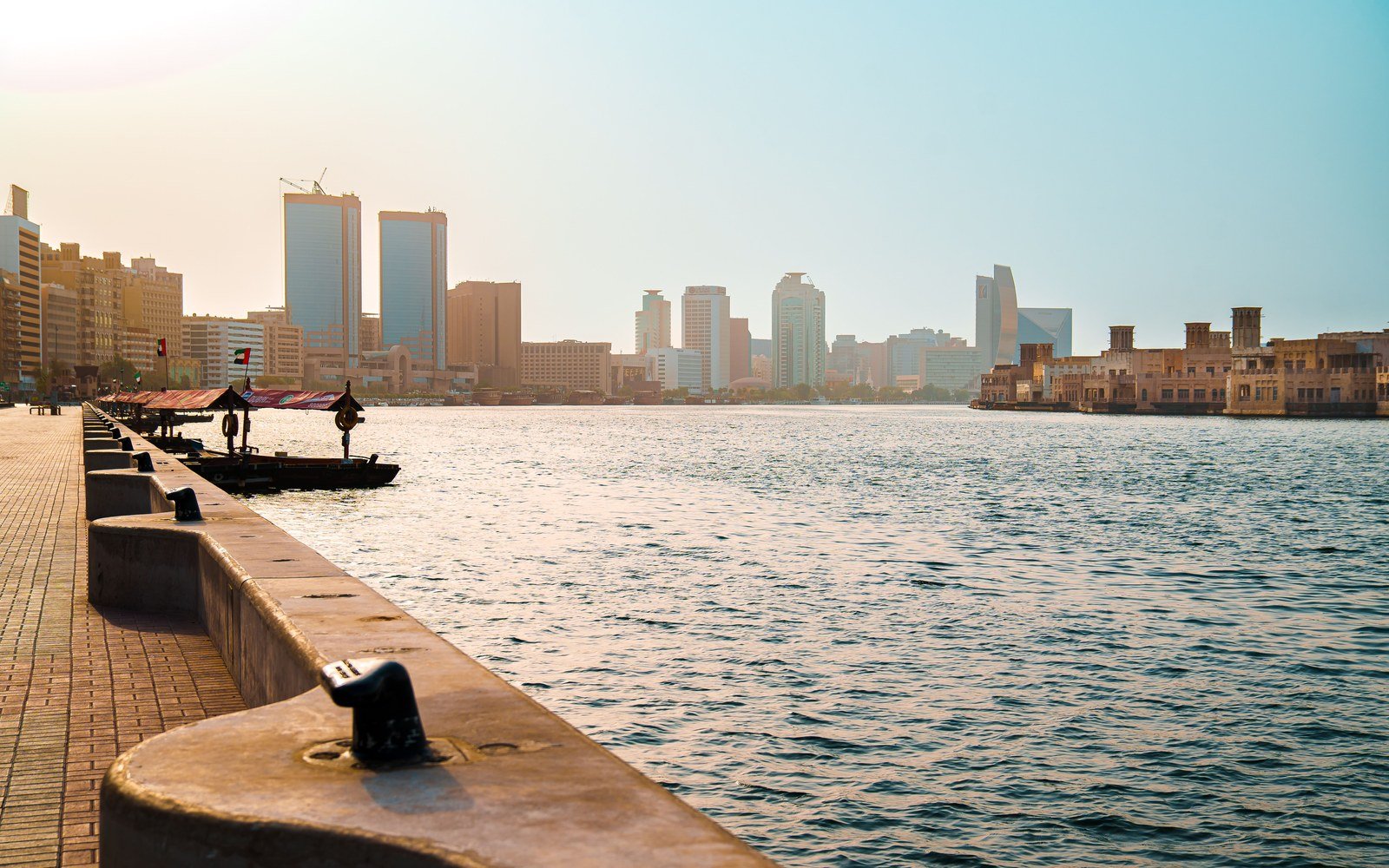 The paved promenade along Dubai Creek in early light, a mooring bollard in the foreground, the water running away to a low skyline of towers and a walled waterfront district on the far bank