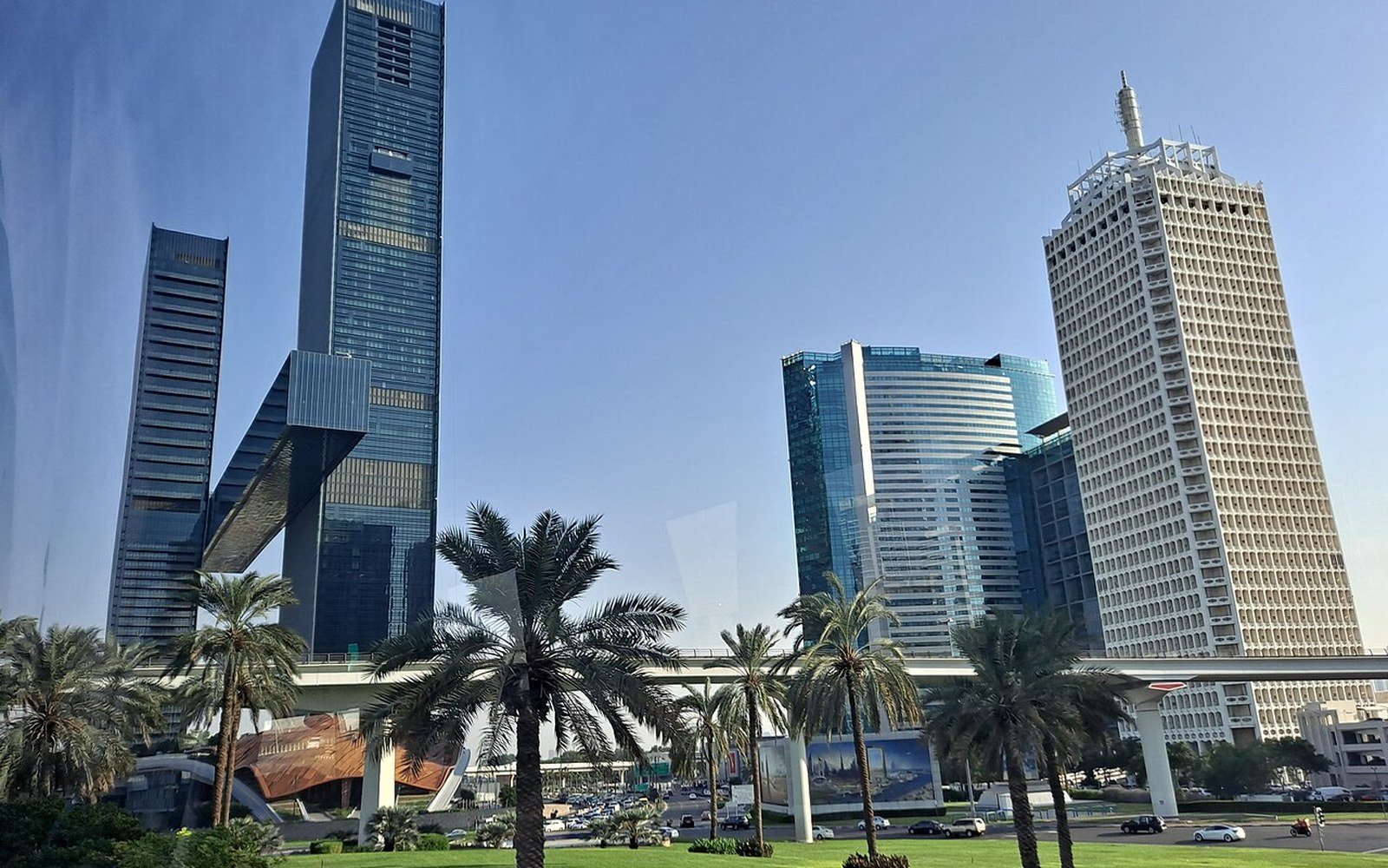 Sheikh Zayed Road in daylight, an elevated railway viaduct crossing the frame in front of office towers, with a row of date palms and cut grass along the roadside in the foreground