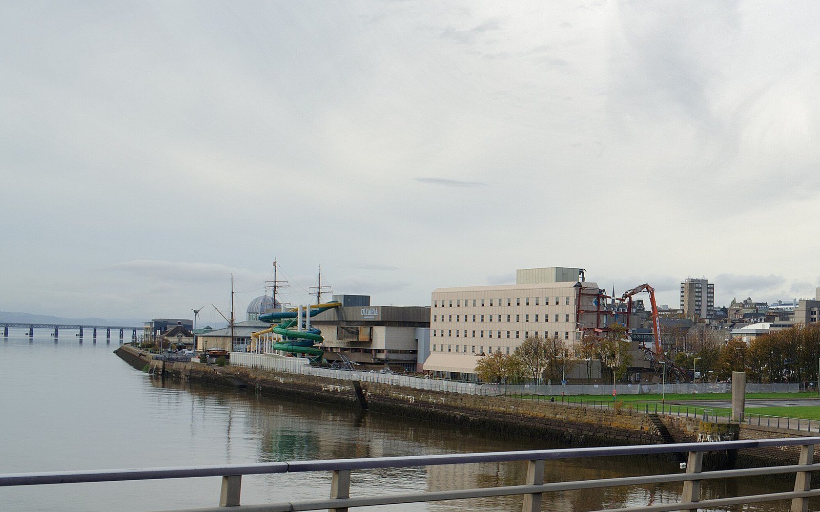 A city waterfront seen across still water, with low quayside buildings, a long road bridge in the haze and grass along the near bank