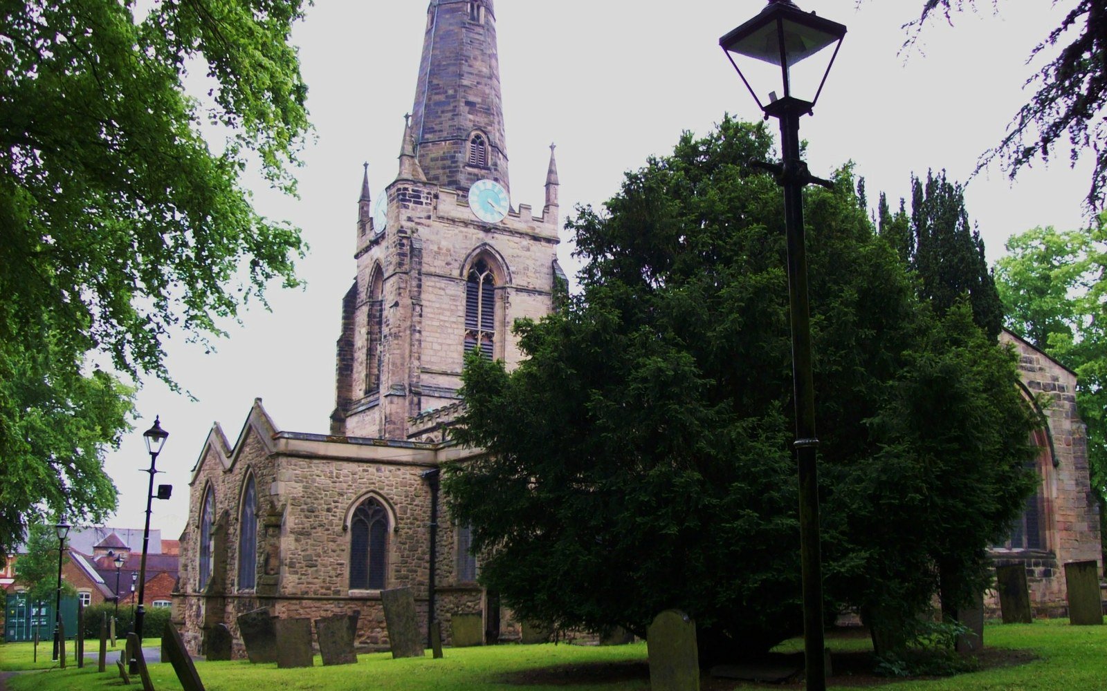 A stone parish church with a tall spire and a clock face, seen across a churchyard through summer trees