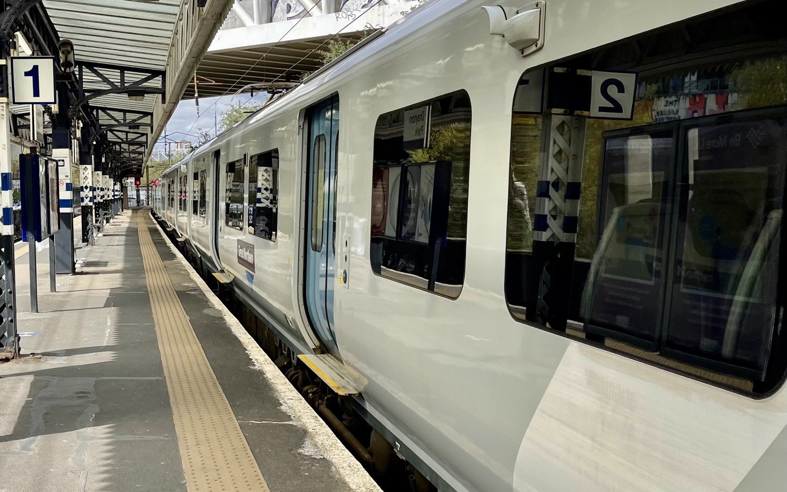 A four car electric passenger train standing at an empty station platform under a canopy, with the running line and overhead wires beyond