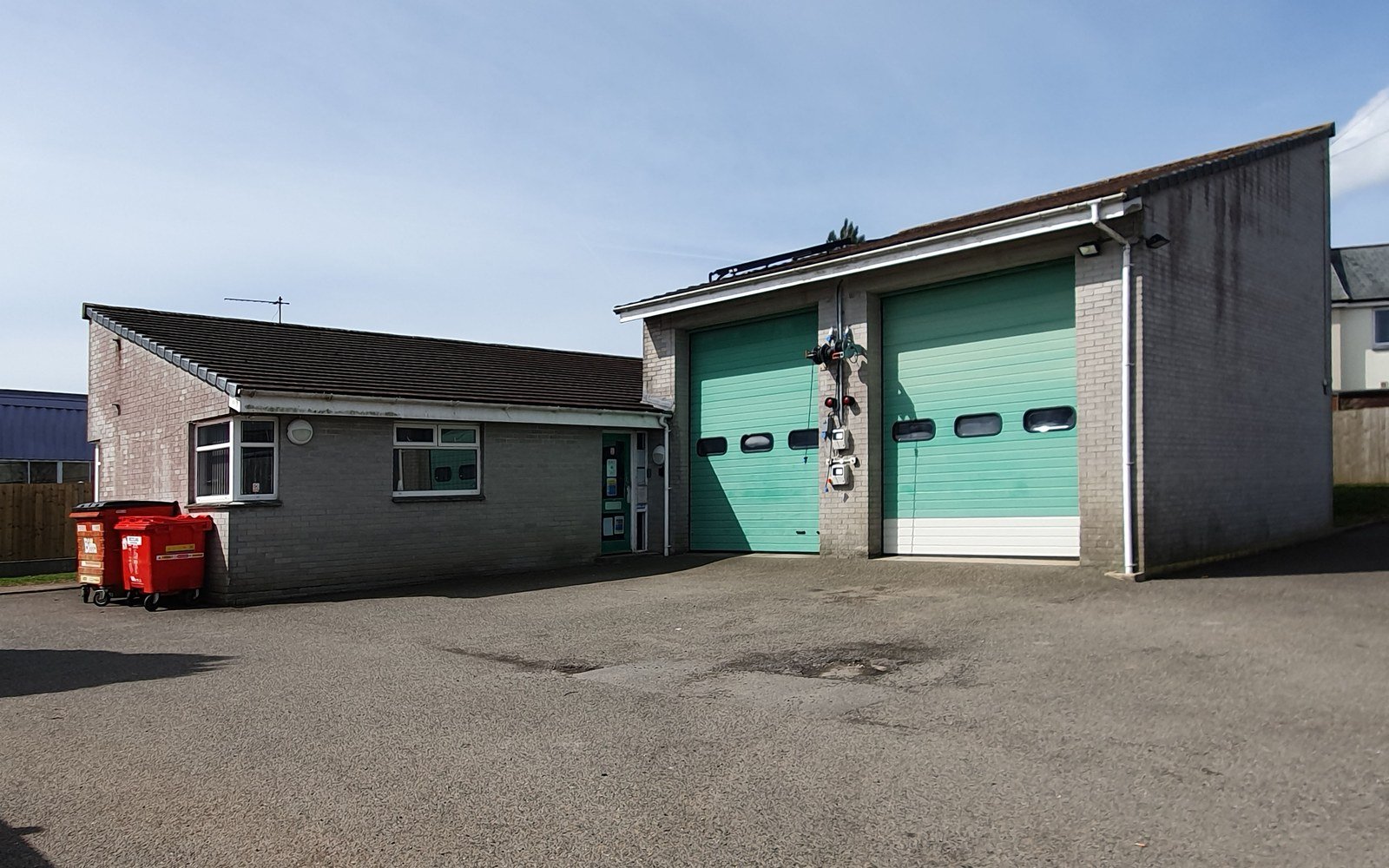 A single storey ambulance station with two wide roller shutter vehicle doors and an adjoining brick office range, seen across an empty apron