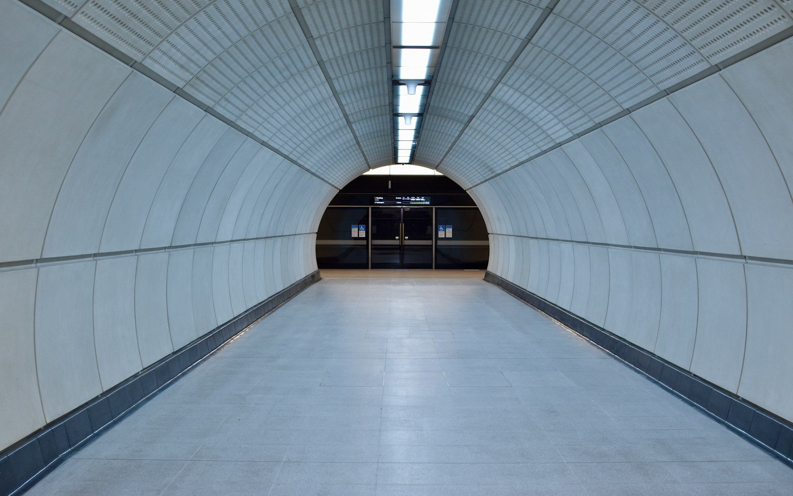A long white-panelled pedestrian tunnel with a strip of ceiling lights leading to a platform entrance