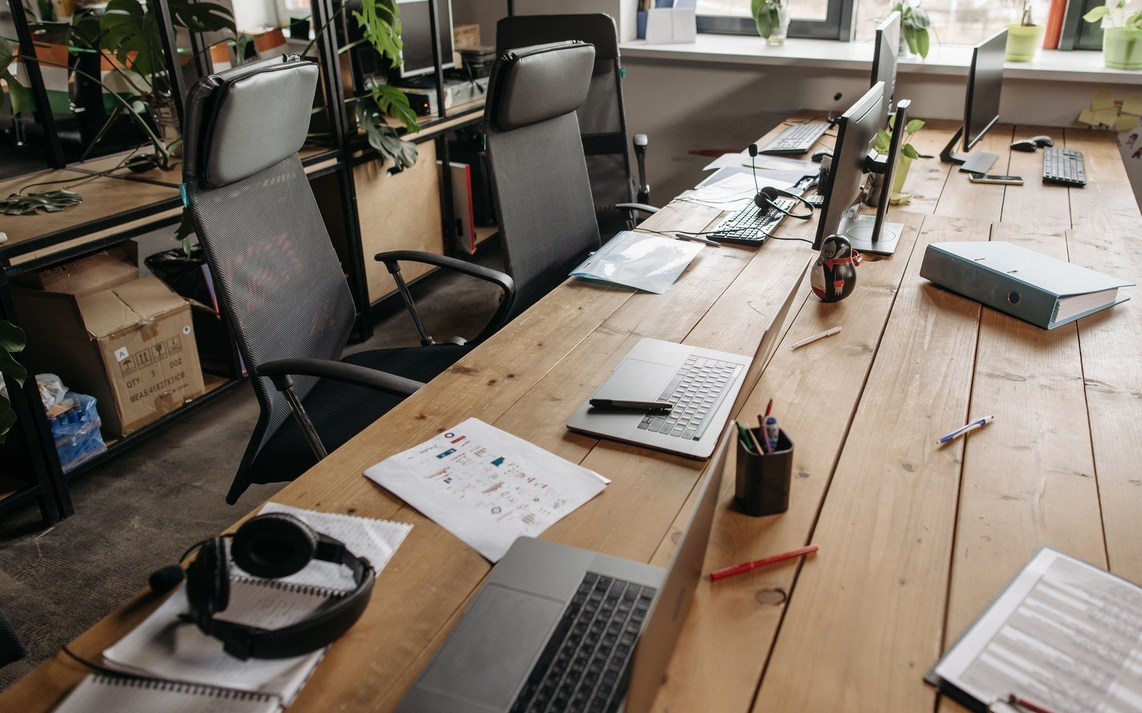 A long shared wooden desk in a workspace, two mesh backed chairs pushed in, open laptops and monitors along the run with notebooks, a printed chart, a pen pot and a ring binder on the boards