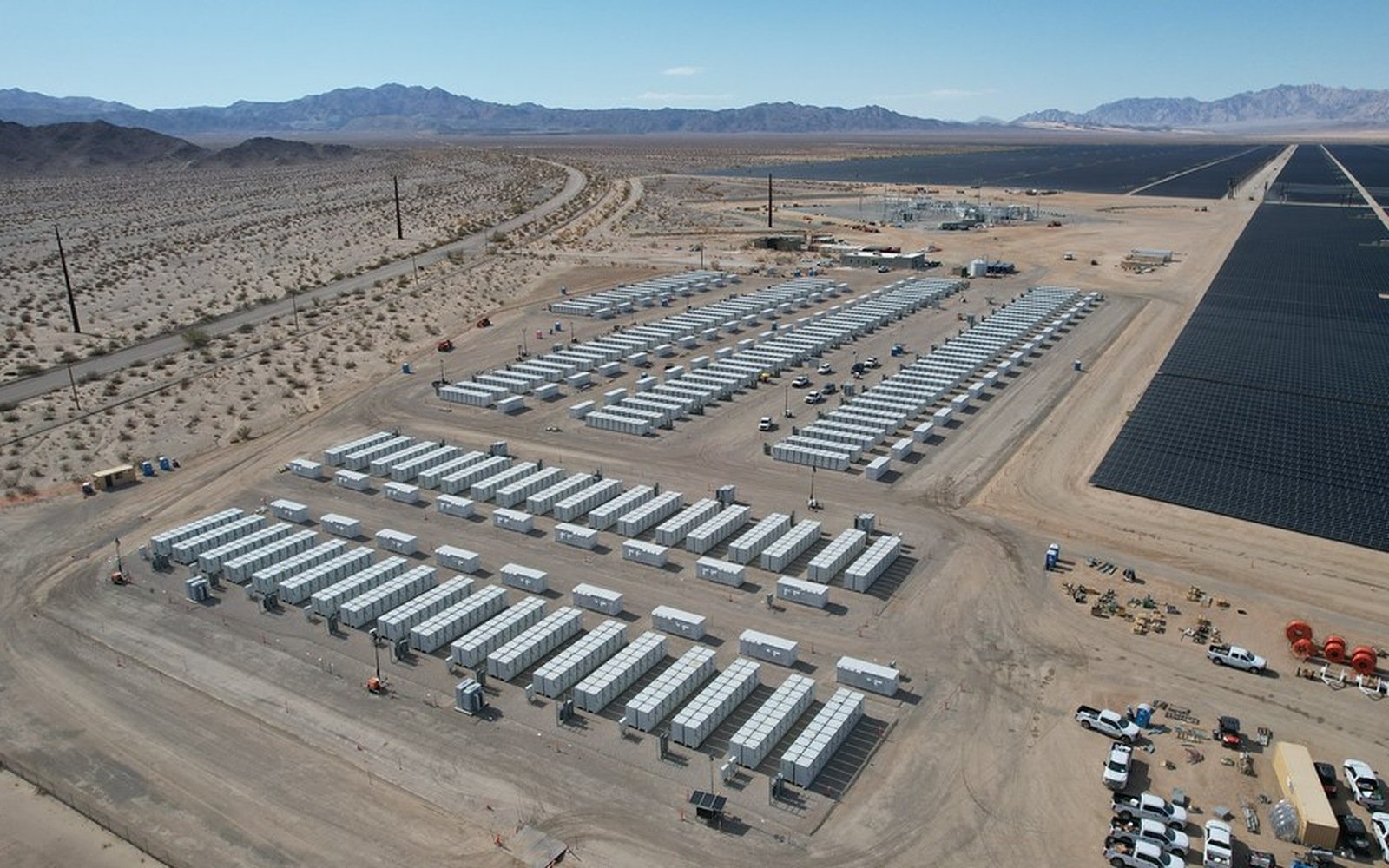 Long parallel rows of white battery containers on a graded desert site, with a solar array on the right hand side and low mountains on the horizon